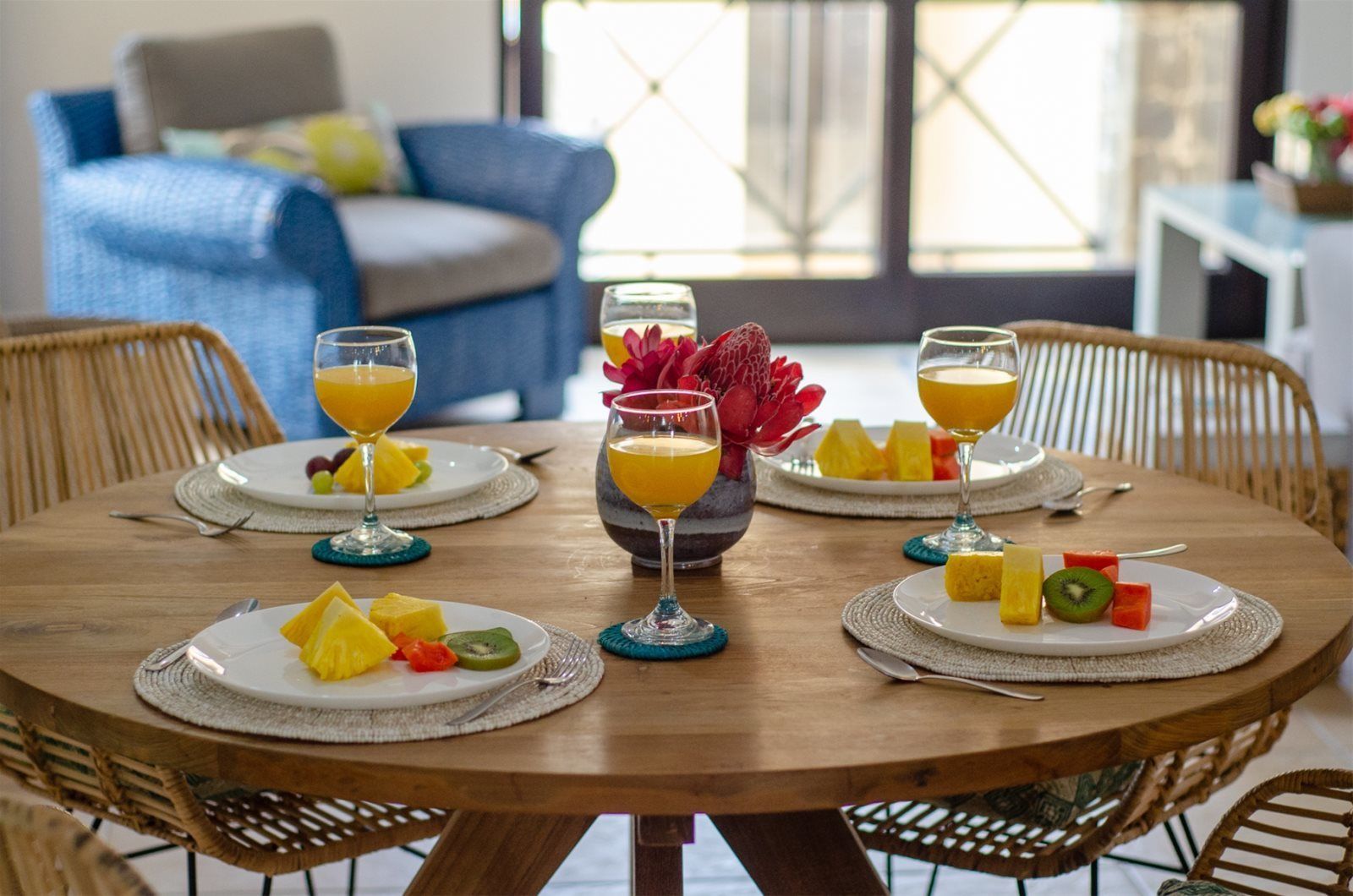 A round wooden dining table with plates of fruit and glasses of orange juice, surrounded by wicker chairs.
