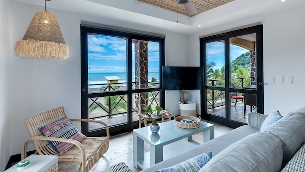 A spacious room with a large glass door leading to a balcony with a view of the ocean. There is a wicker chair, a sofa, and a glass coffee table. - Be-3l (Jacó)