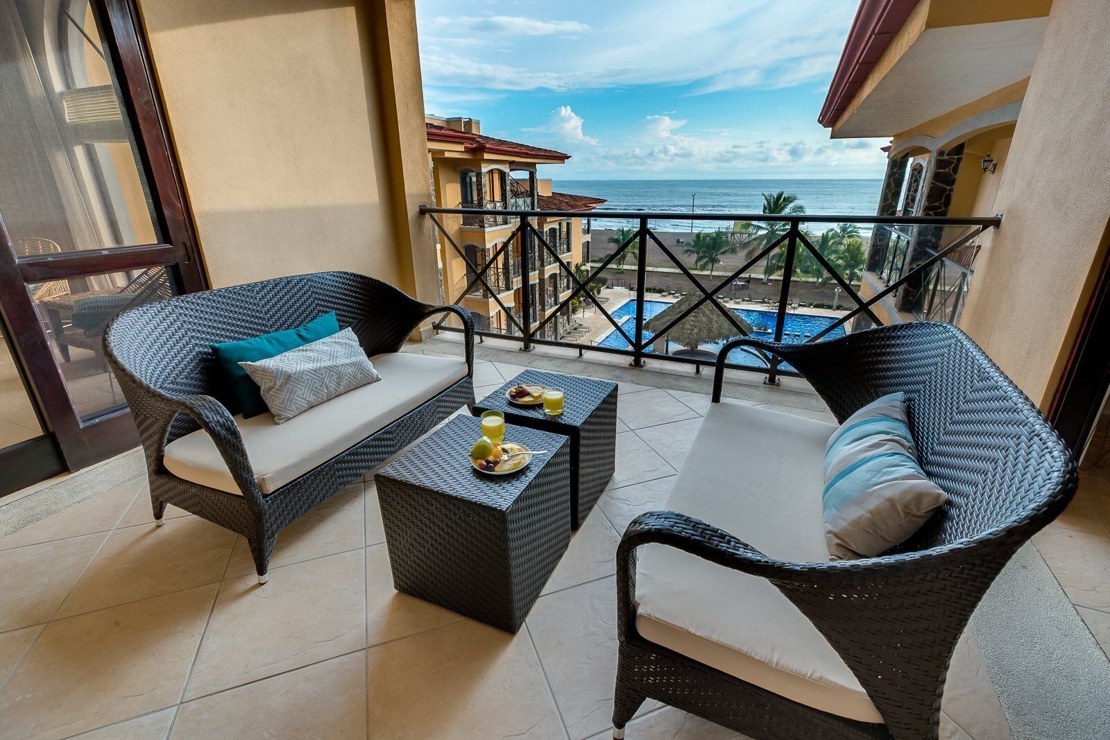 Een balkon met rieten meubels, een tafel met fruit en sap, en uitzicht op de oceaan en gebouwen.