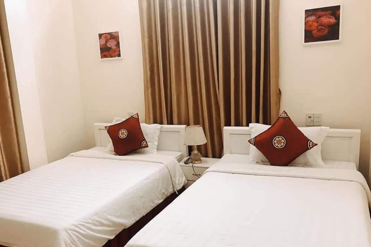 A hotel room with two single beds, each with a white bedspread and a red pillow. There is a large window with curtains, a bedside table with a lamp, and a wall-mounted picture.