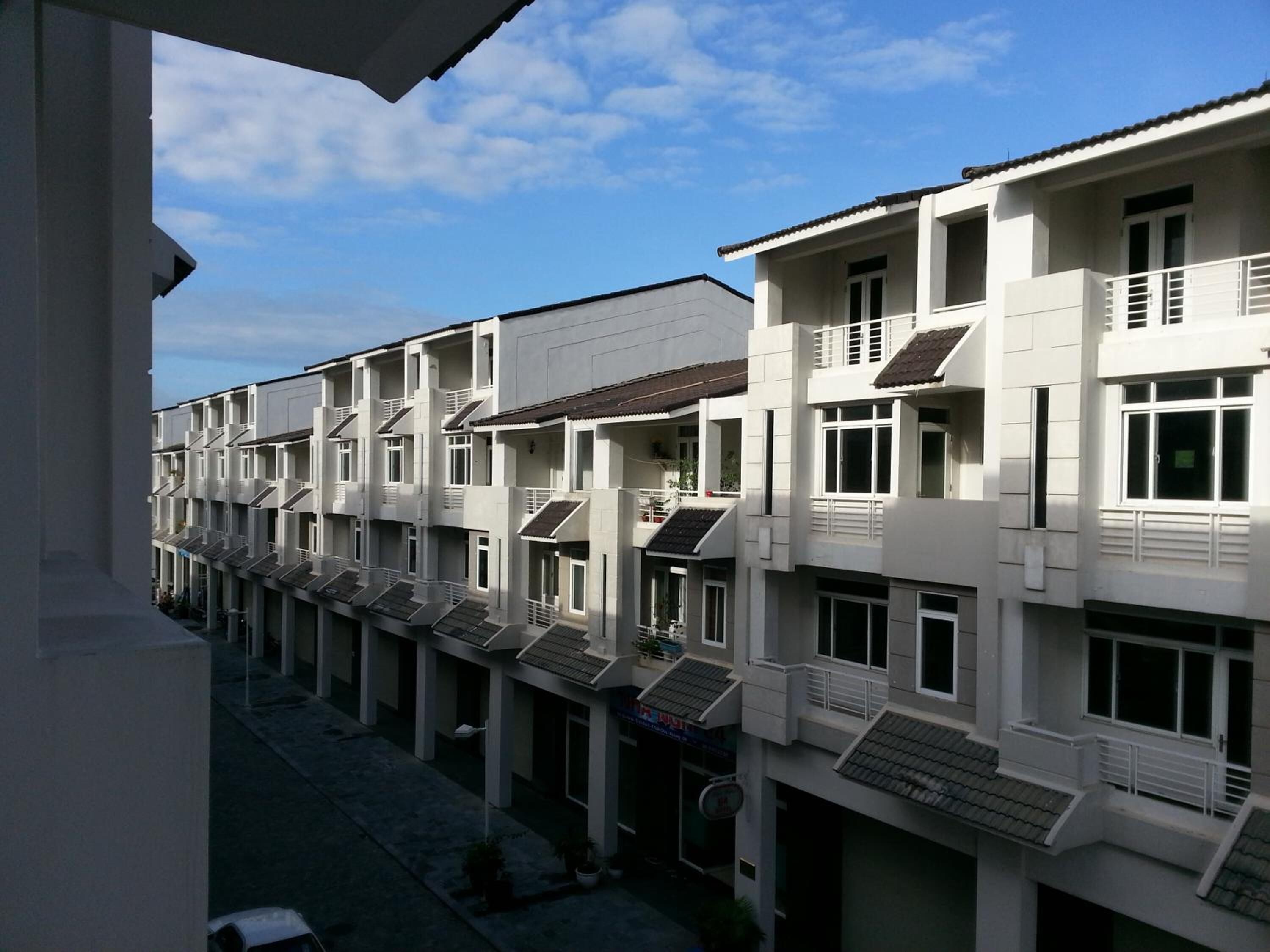 A row of white buildings with balconies, a clear sky, and a paved ground.