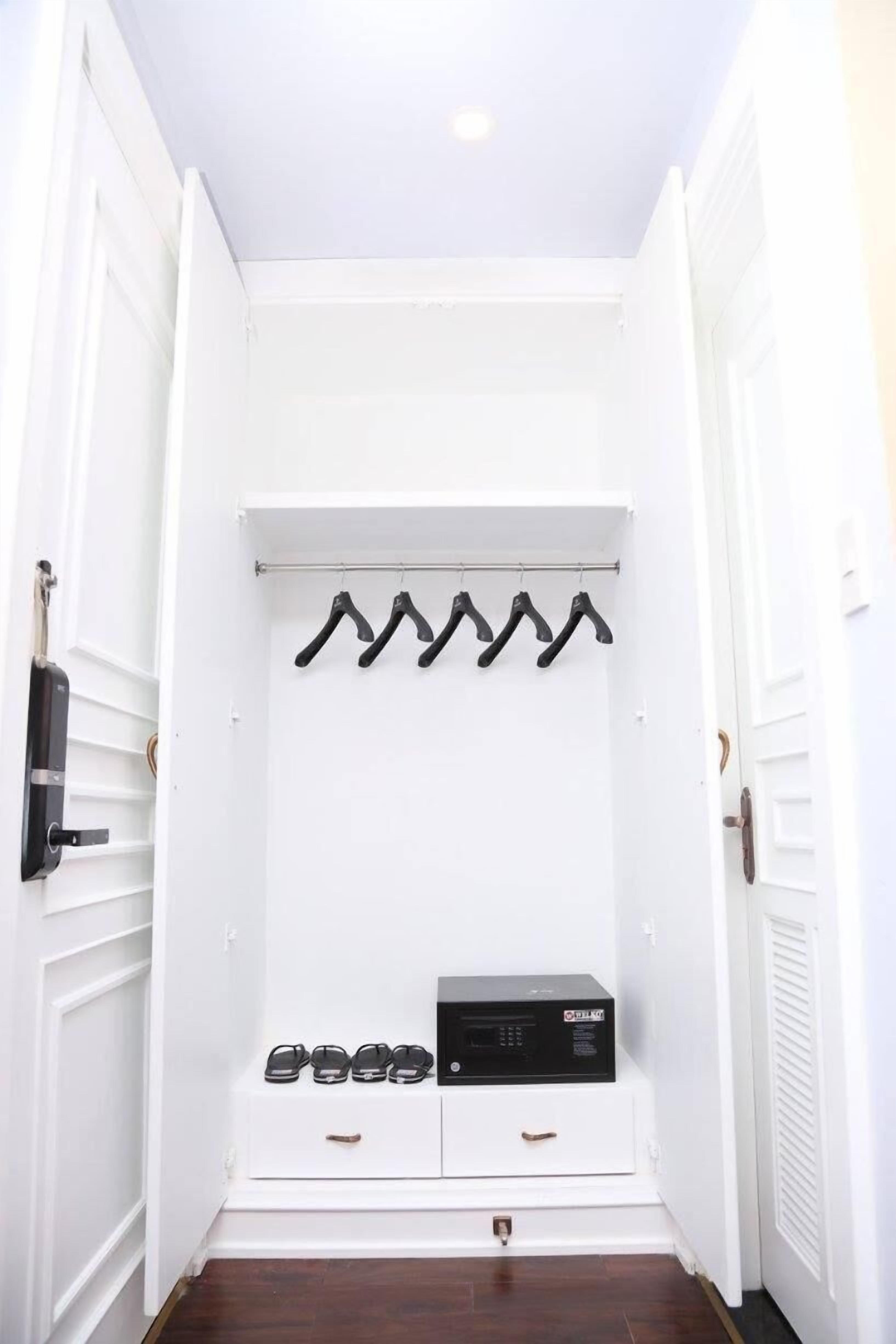 Um corredor com uma porta branca, um porta-roupas com vários cabides, uma pequena prateleira com sapatos e um dispositivo eletrônico preto.