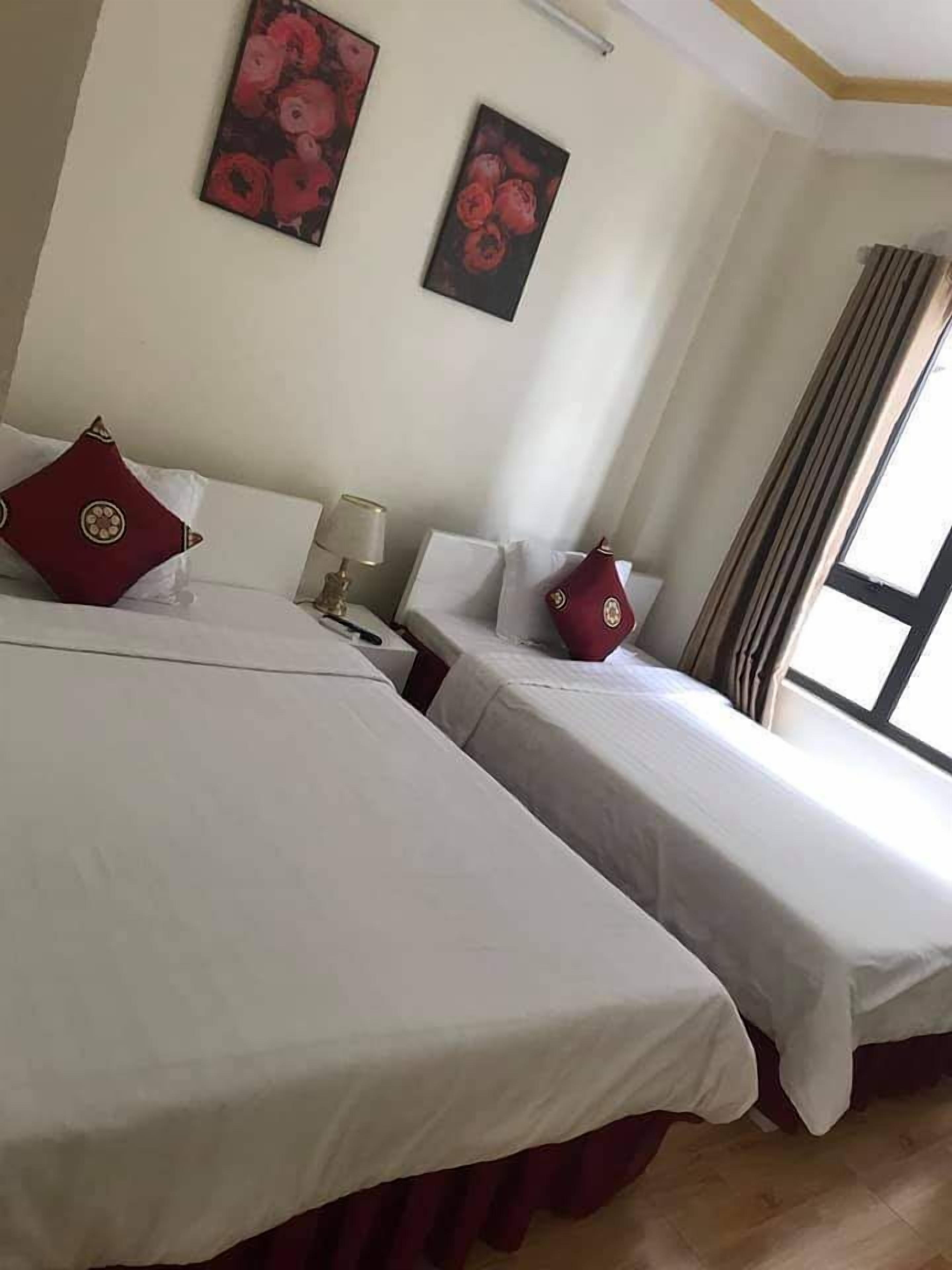 A hotel room with two beds, each with white bedding and red decorative pillows. There are two wall-mounted paintings of flowers, a bedside lamp, and a window with curtains.