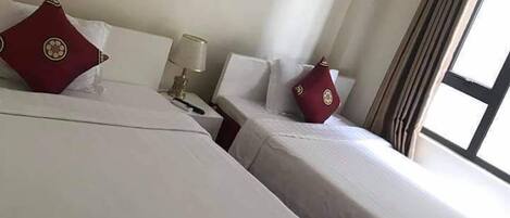 A hotel room with two beds, each with white bedding and red decorative pillows. There are two wall-mounted paintings of flowers, a bedside lamp, and a window with curtains.