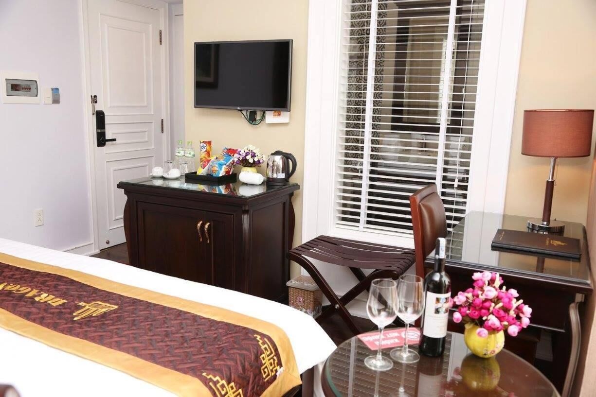 A hotel room with a bed, a television, a desk with a lamp, a window with blinds, and a table with a wine bottle and flowers.
