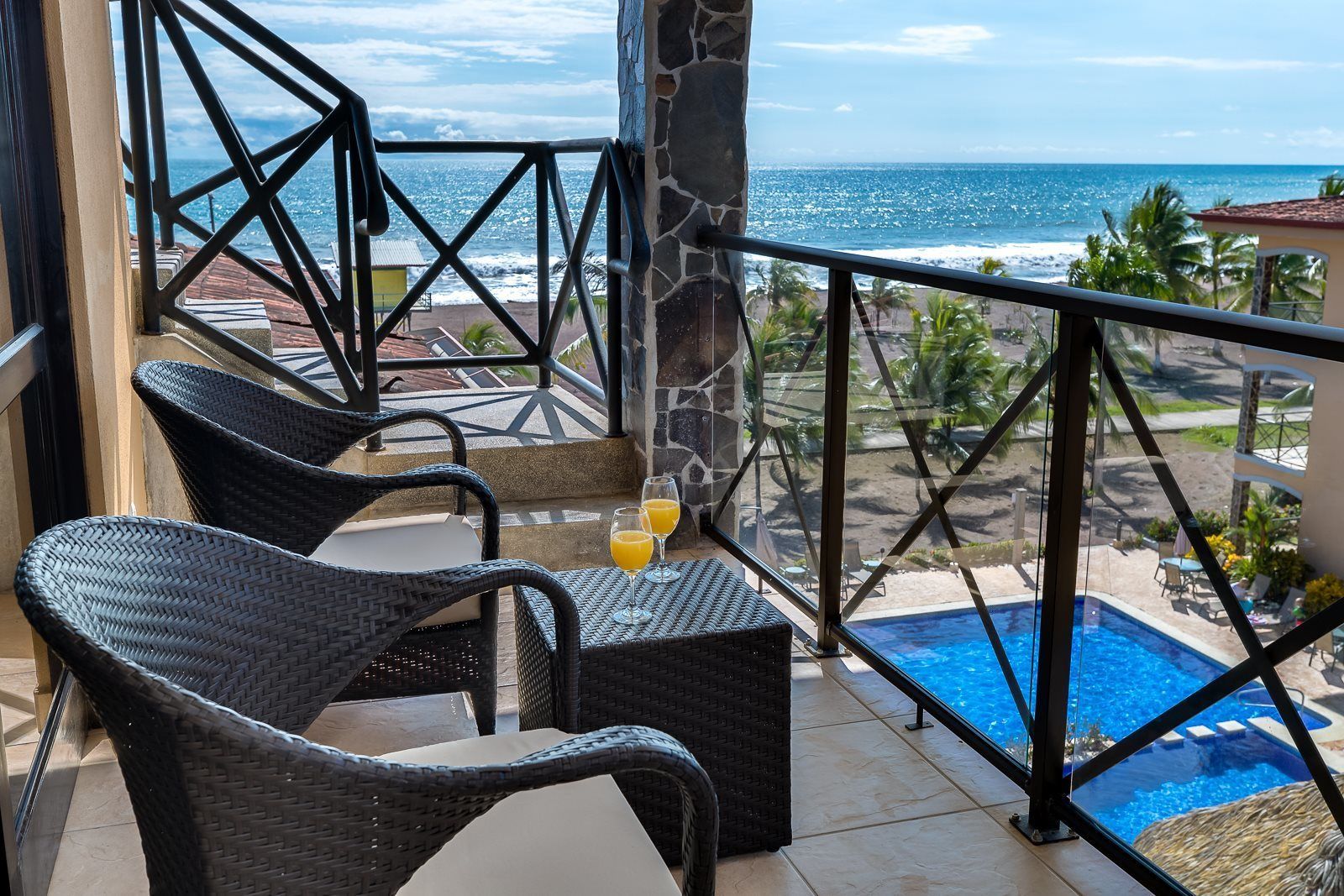 A balcony with wicker chairs, a table, and a glass of orange juice. A pool is visible in the background.
