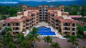 A multi-story hotel with a central swimming pool, surrounded by palm trees and a mountainous backdrop. - Perfect Oceanfront Retreat for Your Family (Jacó)