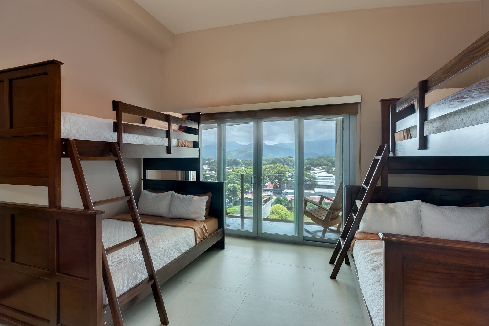 A dormitory room with bunk beds, a large window with a view of a cityscape, and a wooden wardrobe.