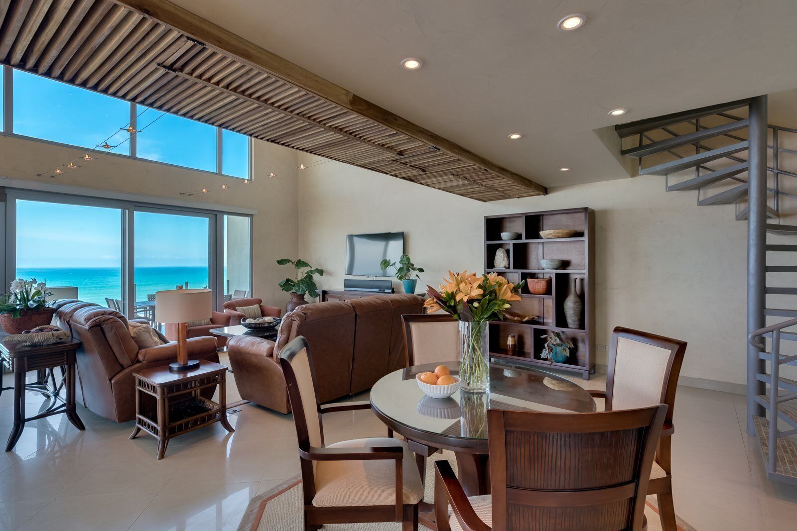 A spacious living area with a large window overlooking the ocean, a dining table with chairs, a sofa, and a bookshelf.