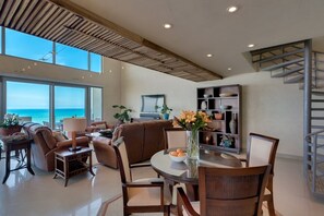 Amplia sala de estar con un gran ventanal con vistas al océano, una mesa de comedor con sillas, un sofá y una estantería.