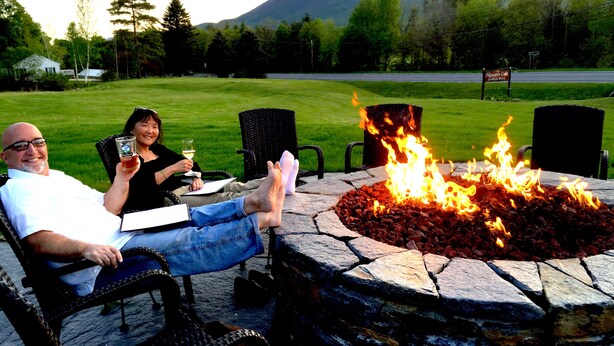 Two people sitting by a stone fire pit with a grassy field and mountains in the background. - Stanford House (Manchester Center)