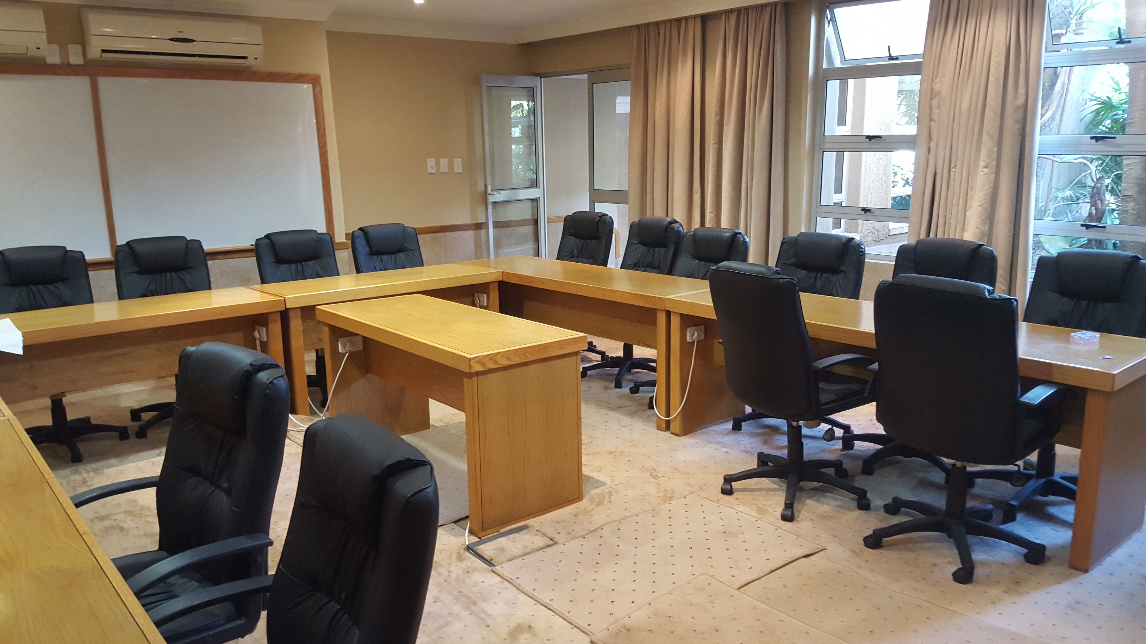 A conference room with a long wooden table, black office chairs, a whiteboard, and a window with curtains.