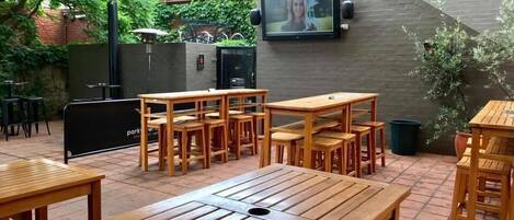 An outdoor patio with wooden tables and chairs, a TV mounted on the wall, and a brick wall with greenery.