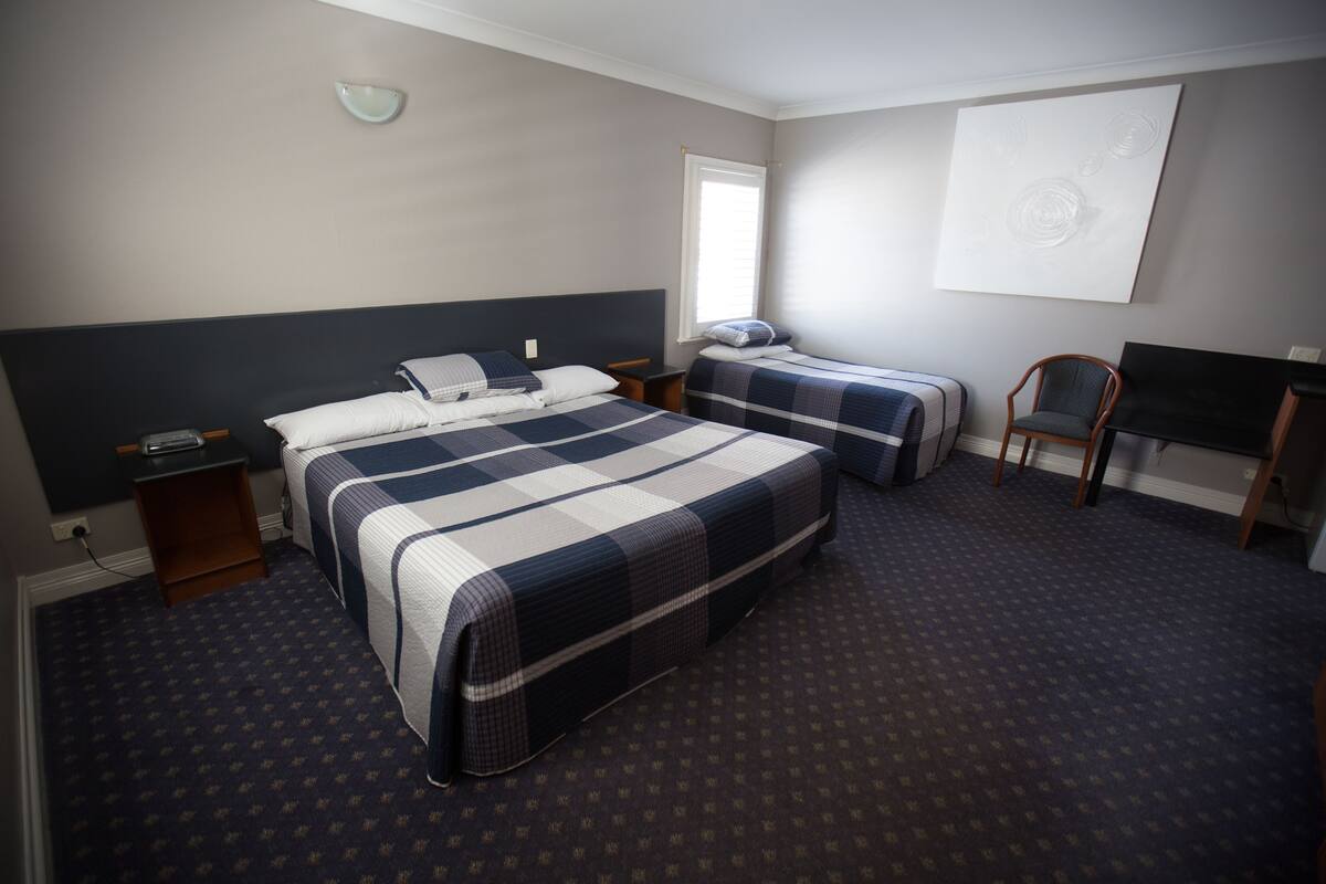 Ein Hotelzimmer mit einem großen Bett, einem kleineren Bett, einem Schreibtisch, einem Sessel, einem Fernseher und einem Fenster.