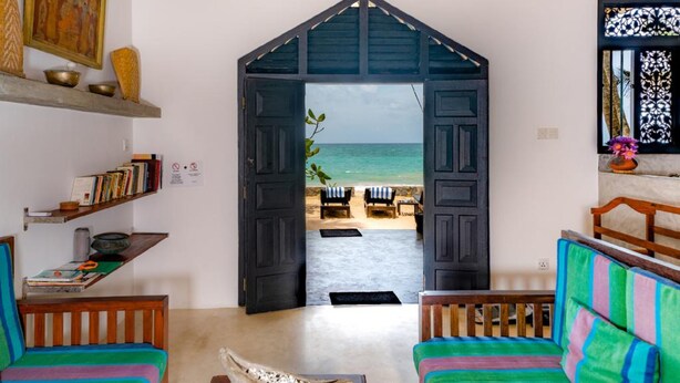 Una habitación con vistas a la playa a través de una puerta abierta, una estantería, un banco de madera y un sofá colorido.