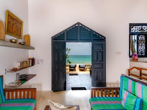 Ein Zimmer mit Blick auf den Strand durch eine geöffnete Tür, ein Bücherregal, eine hölzerne Bank und ein farbenfrohes Sofa.