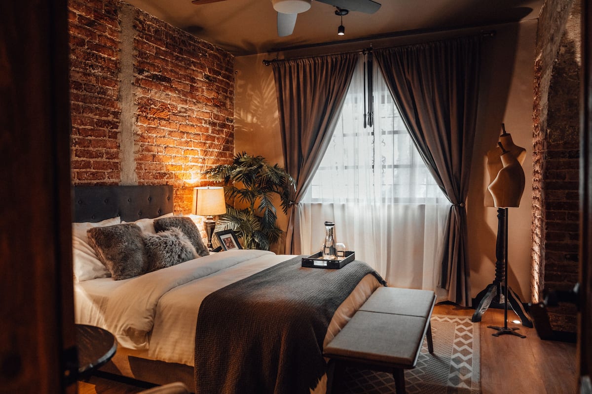 Ein Schlafzimmer mit Backsteinwand, einem Bett mit Kopfteil, einem Nachttisch mit Lampe, einer Bank und einem Fenster mit Vorhängen.
