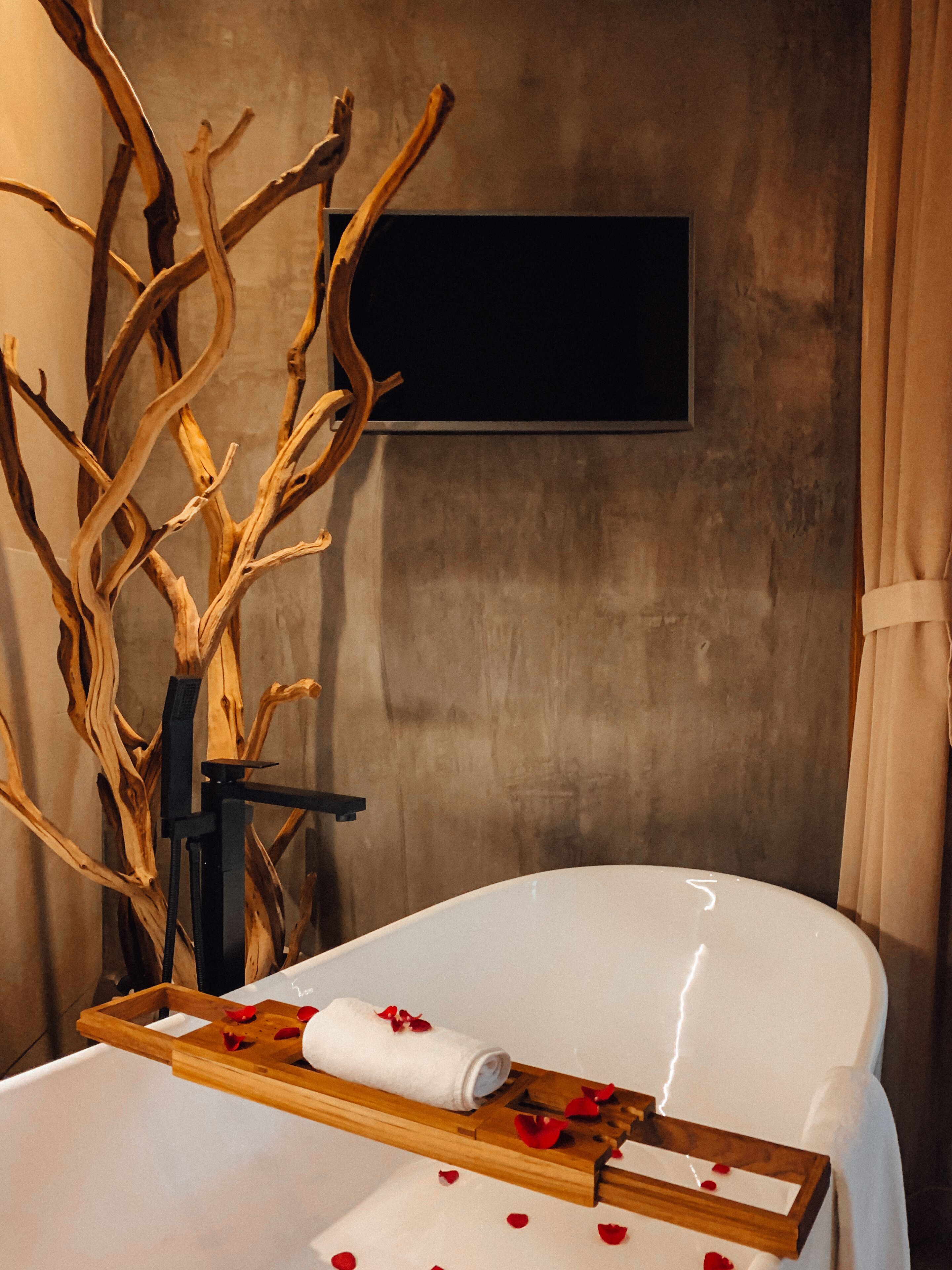 Une salle de spa avec un fauteuil blanc, un plateau en bois sur lequel sont posés une serviette roulée et des pétales de rose, ainsi qu’une grande branche d’arbre utilisée comme élément de décoration.