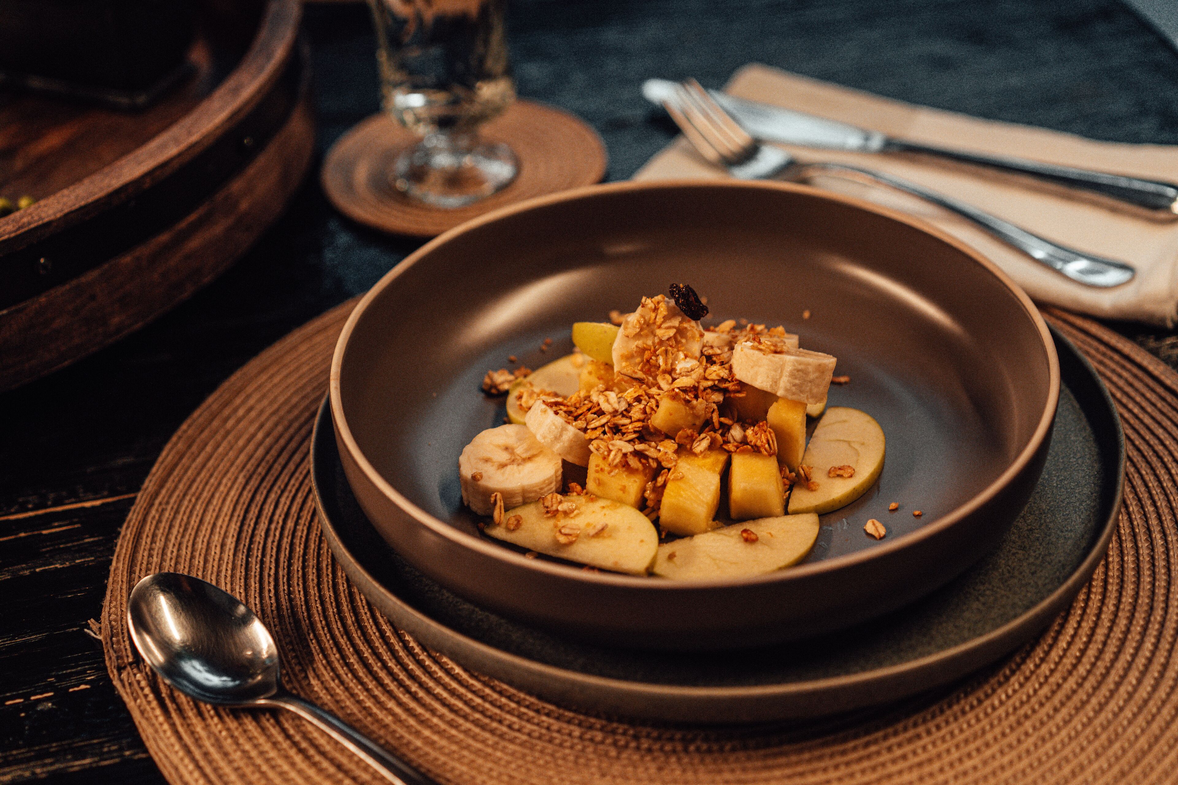 Un tazón de rodajas de plátano cubierto de granola y nueces sobre un plato oscuro, junto a una cuchara y un vaso de agua sobre un tapete tejido.