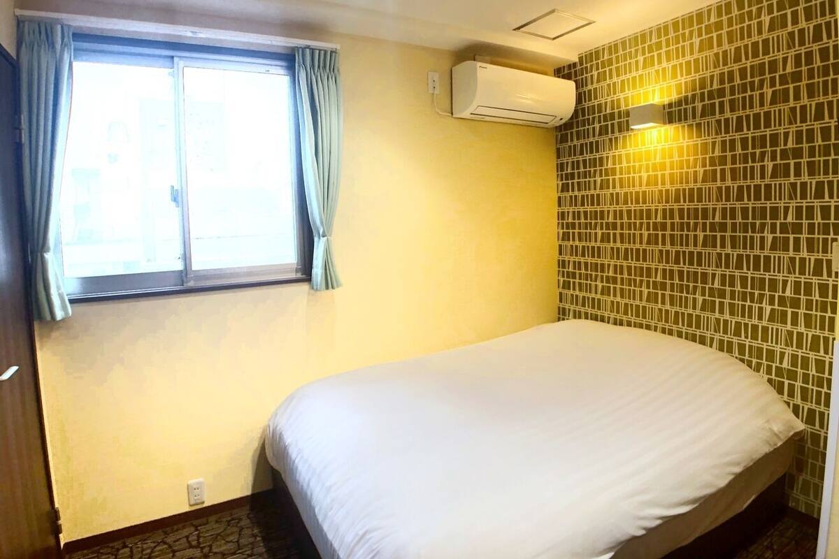 Une chambre avec un lit, une fenêtre avec des rideaux, un appareil de climatisation et un mur orné d’un motif géométrique.