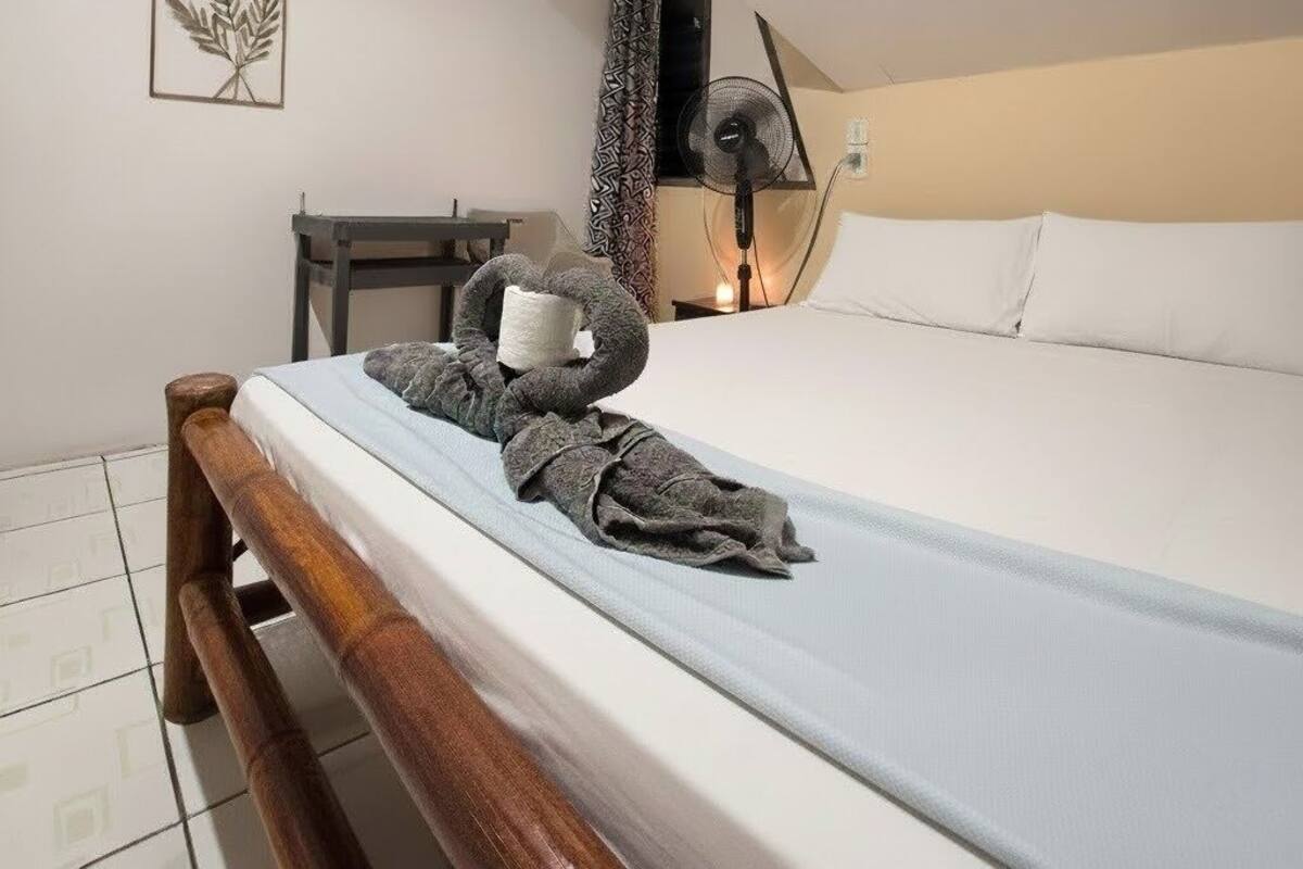 A neatly made bed with a grey blanket and a rolled towel, a bedside table with a lamp, and a wall-mounted fan.