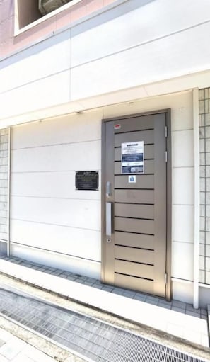 A closed metal door with a sign, a small plaque, and a metal grate on the ground. - ABC Guesthouse 1F (Izumisano)