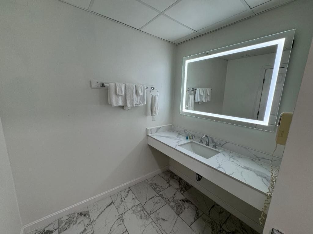 Un baño con lavamanos de mármol blanco, un espejo grande con iluminación integrada y un perchero con varias toallas.