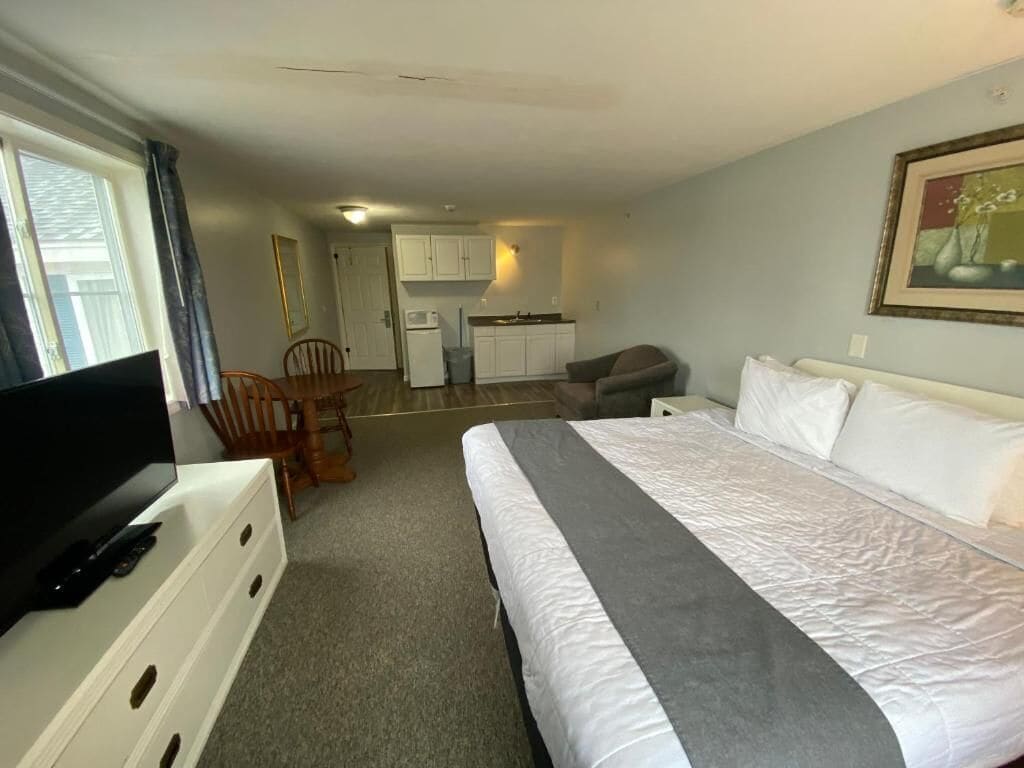 A hotel room with a bed, a TV, a desk, and a kitchenette in the background.