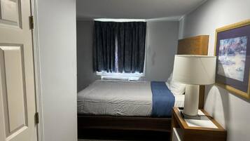 Un dormitorio con cama, mesita de noche con lámpara, ventana con cortinas y un cuadro en la pared.