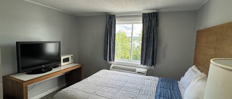 A hotel room with a bed, a TV, a small table, a window with curtains, and a view of greenery outside.