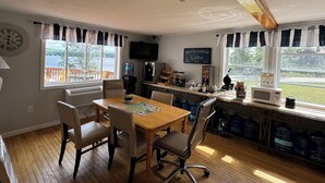 A dining area with a wooden table and chairs, a clock on the wall, and a view of a lake. - Winnisquam Lake Inn (Tilton)