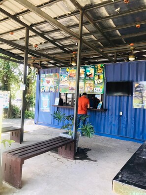 Un espacio de estar al aire libre con techo, una barra, una persona en la barra y un contenedor azul con tarifas e información sobre servicios.