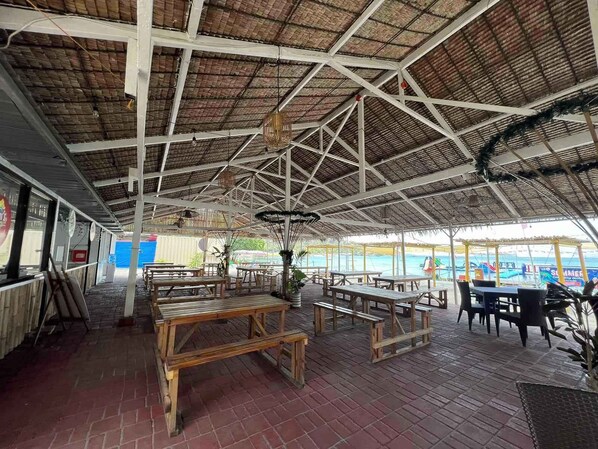 Un espacio de patio al aire libre con mesas y bancos de madera, techo de paja y vista al mar.