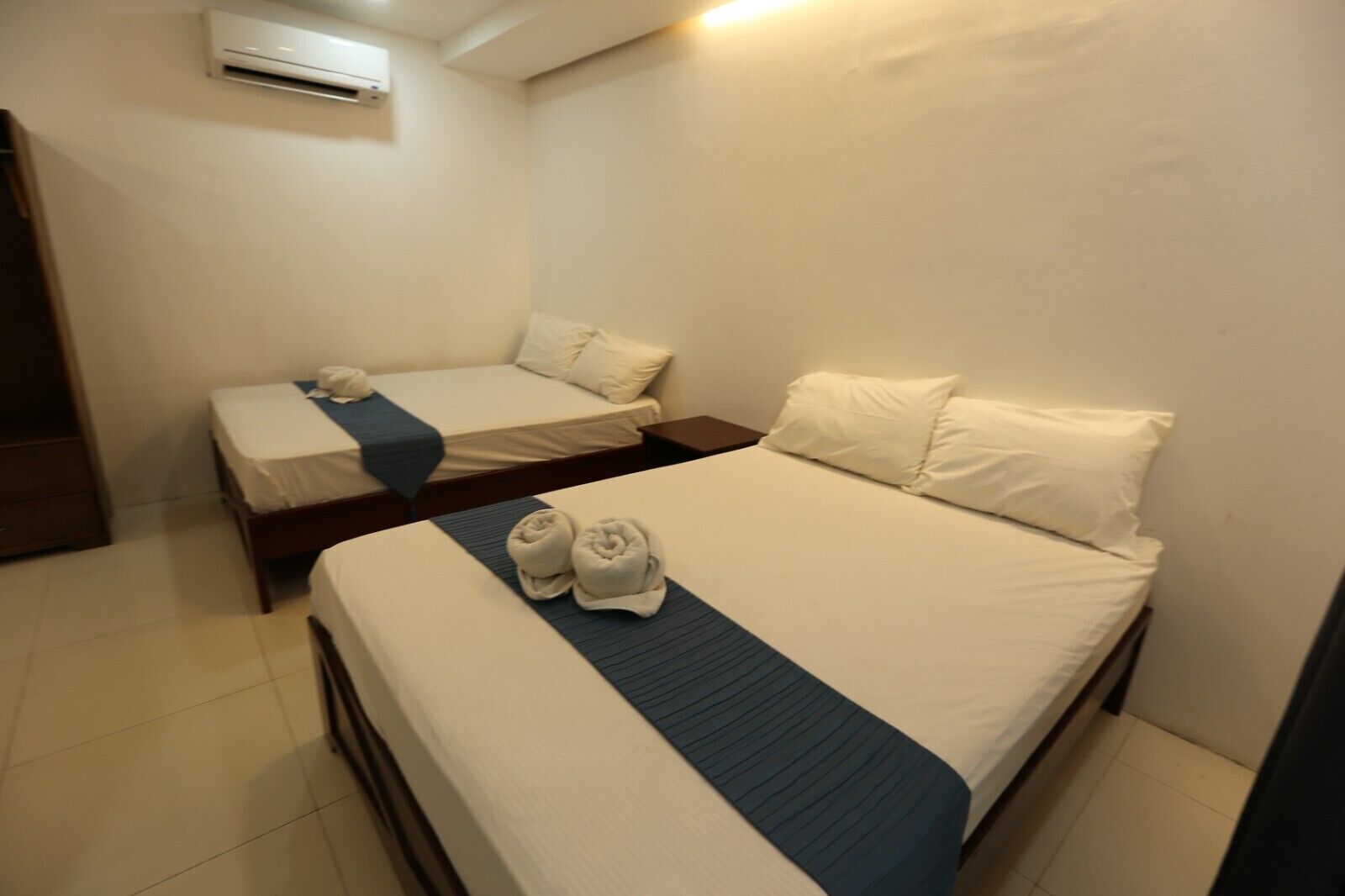 A hotel room with two beds, a wooden nightstand, and an air conditioning unit on the wall.