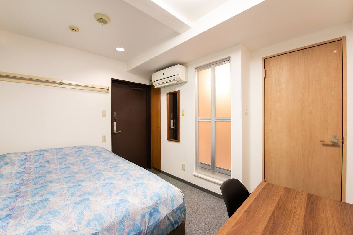 A room with a bed, a wooden desk, a window, and an air conditioning unit.