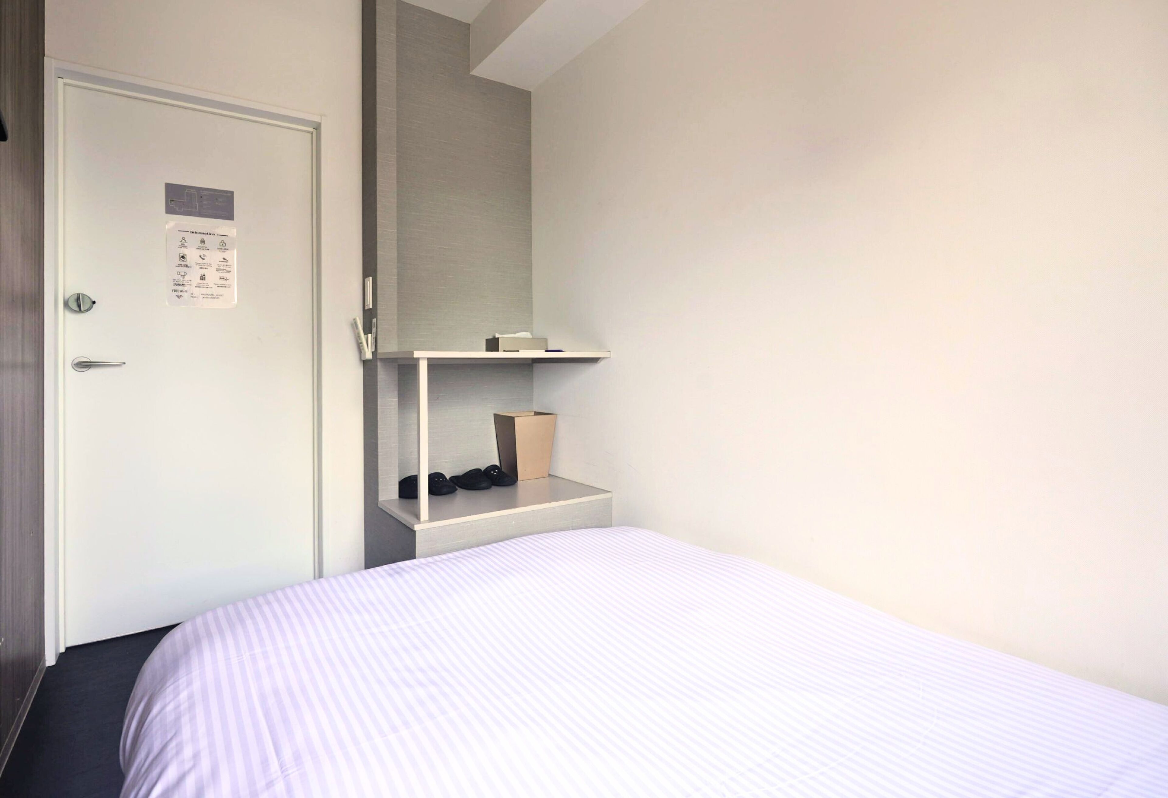A bedroom with a bed, a door, a small shelf with shoes, and a wall-mounted shelf.