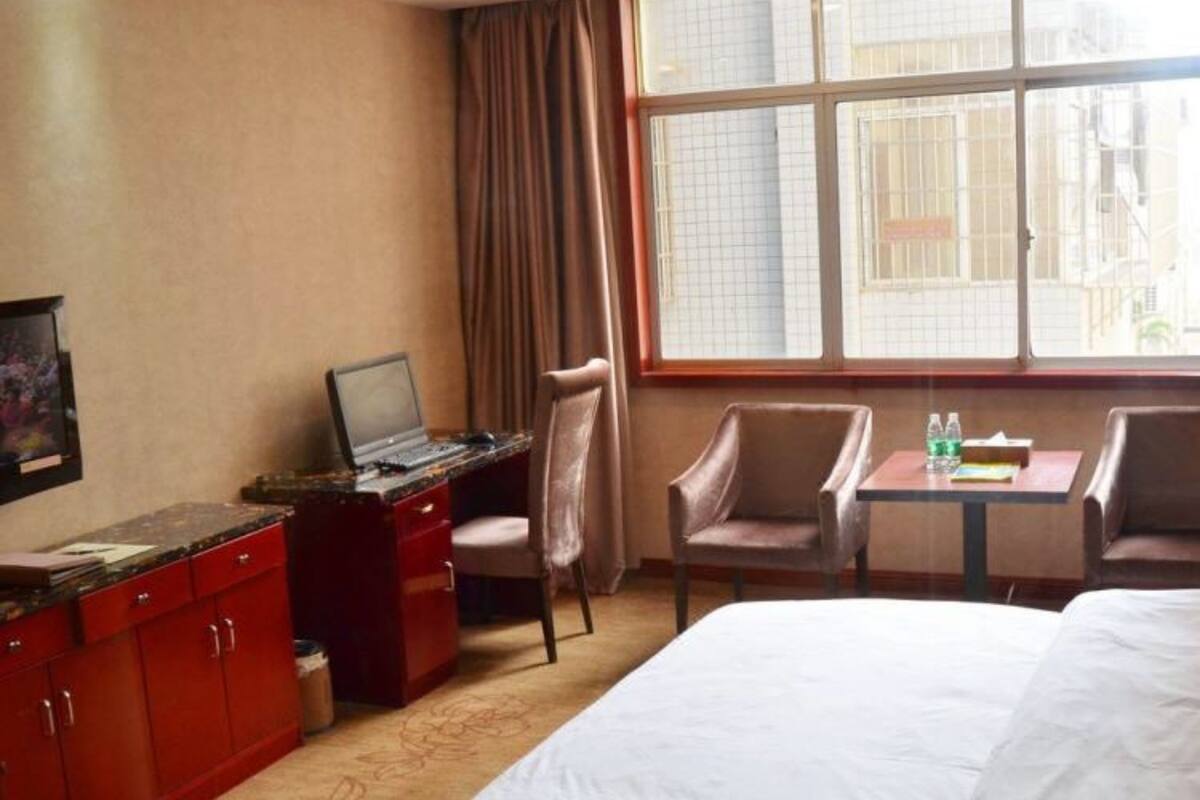 Ein Hotelzimmer mit einem Bett, einem Schreibtisch mit einem Laptop, zwei Stühlen, einem kleinen Tisch und einem Fenster mit Vorhängen.