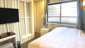 Ett hotellrum med en säng, ett skrivbord, en TV, en garderob och ett fönster med persienner.