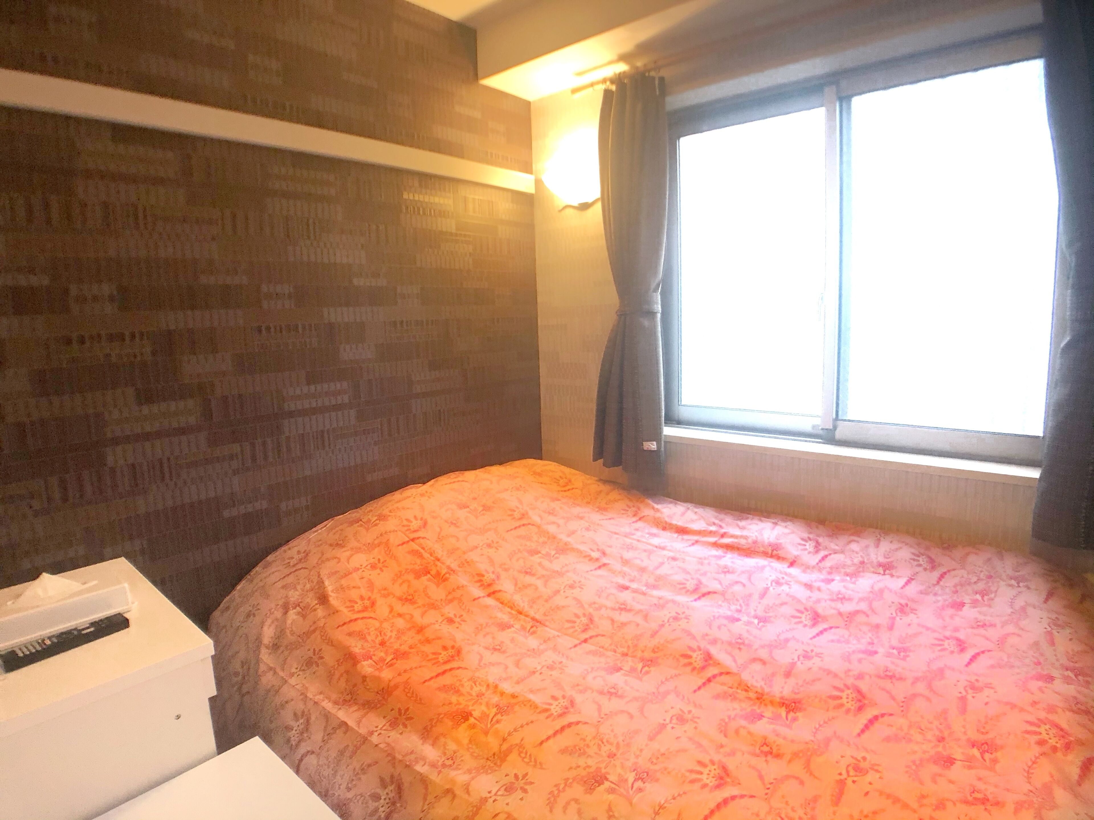 A bedroom with a patterned bedspread, a bedside table with a tissue box, a window with curtains, and a brick-patterned wall.