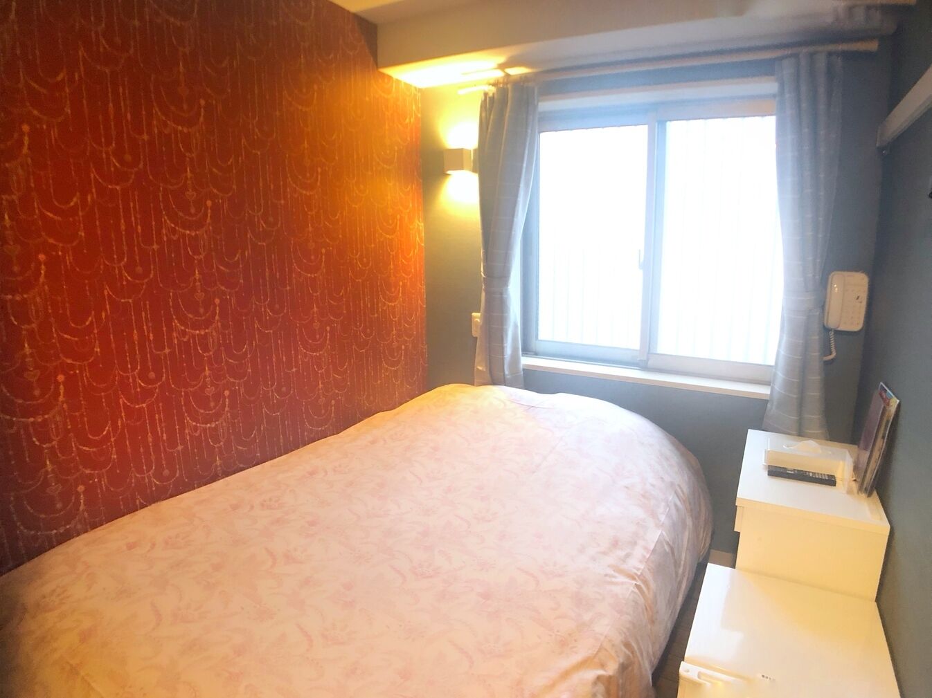 A single bed in a room with a patterned wall, a window with curtains, and a bedside table with a phone and a remote control.