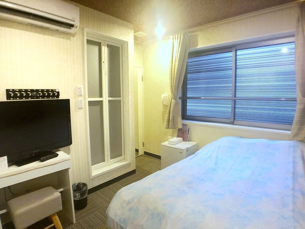A hotel room with a bed, a TV, a desk, a window with blinds, and a door.