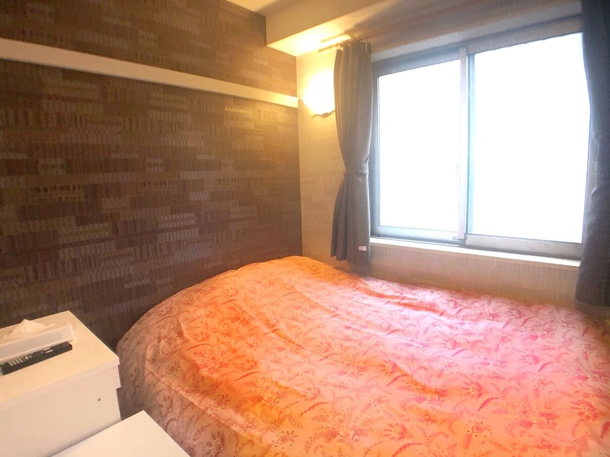 A room with a bed, a window with curtains, a wall with a patterned design, and a white object on the floor.
