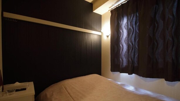 Um quarto pequeno, com pouca luz, com uma cama de solteiro, uma mesa de cabeceira e uma luminária na parede.