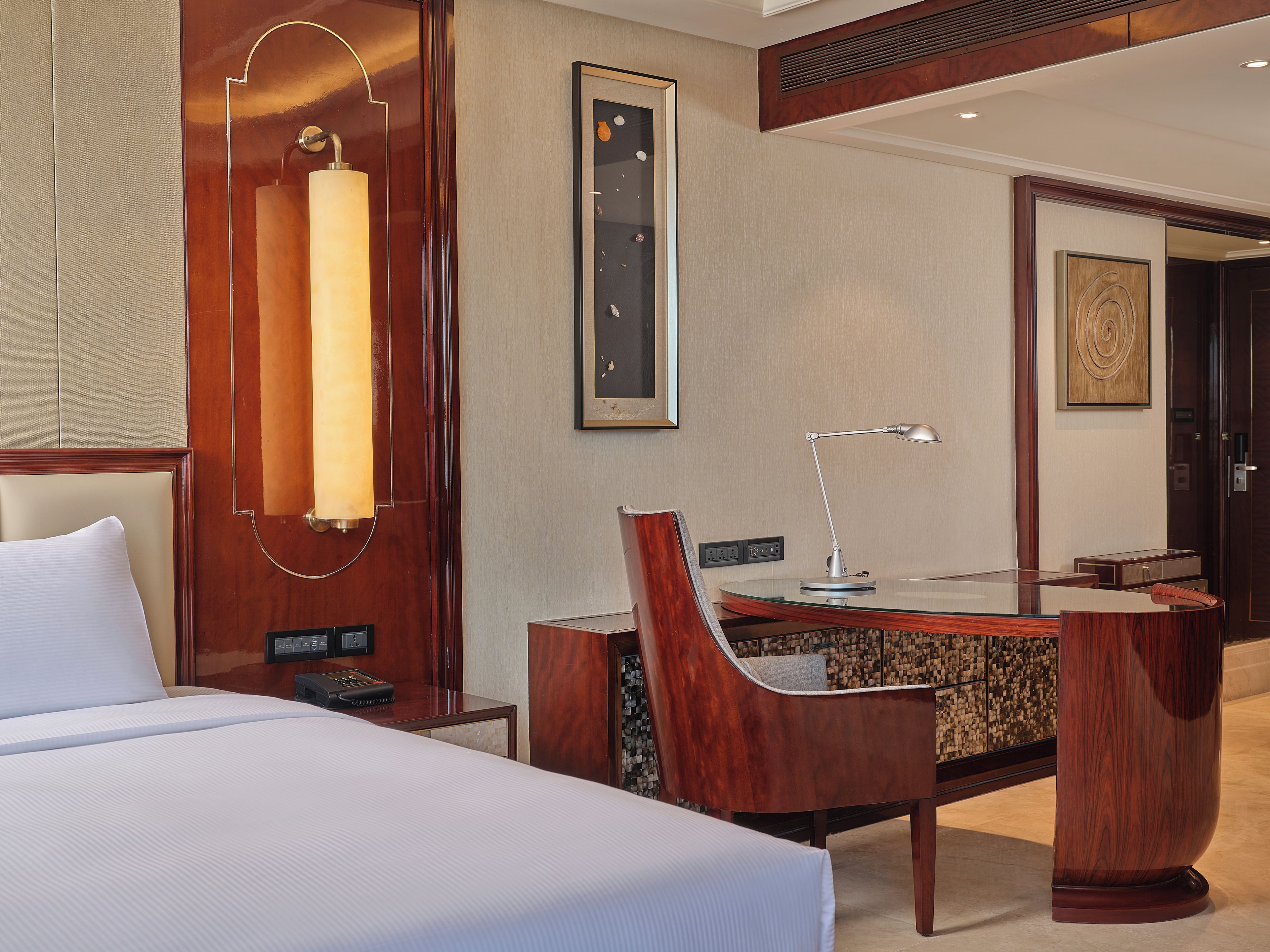 A hotel room with a bed, a desk with a chair, a desk lamp, a telephone, and a wall-mounted artwork.