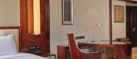 A hotel room with a bed, a desk with a chair, a desk lamp, a telephone, and a wall-mounted artwork.