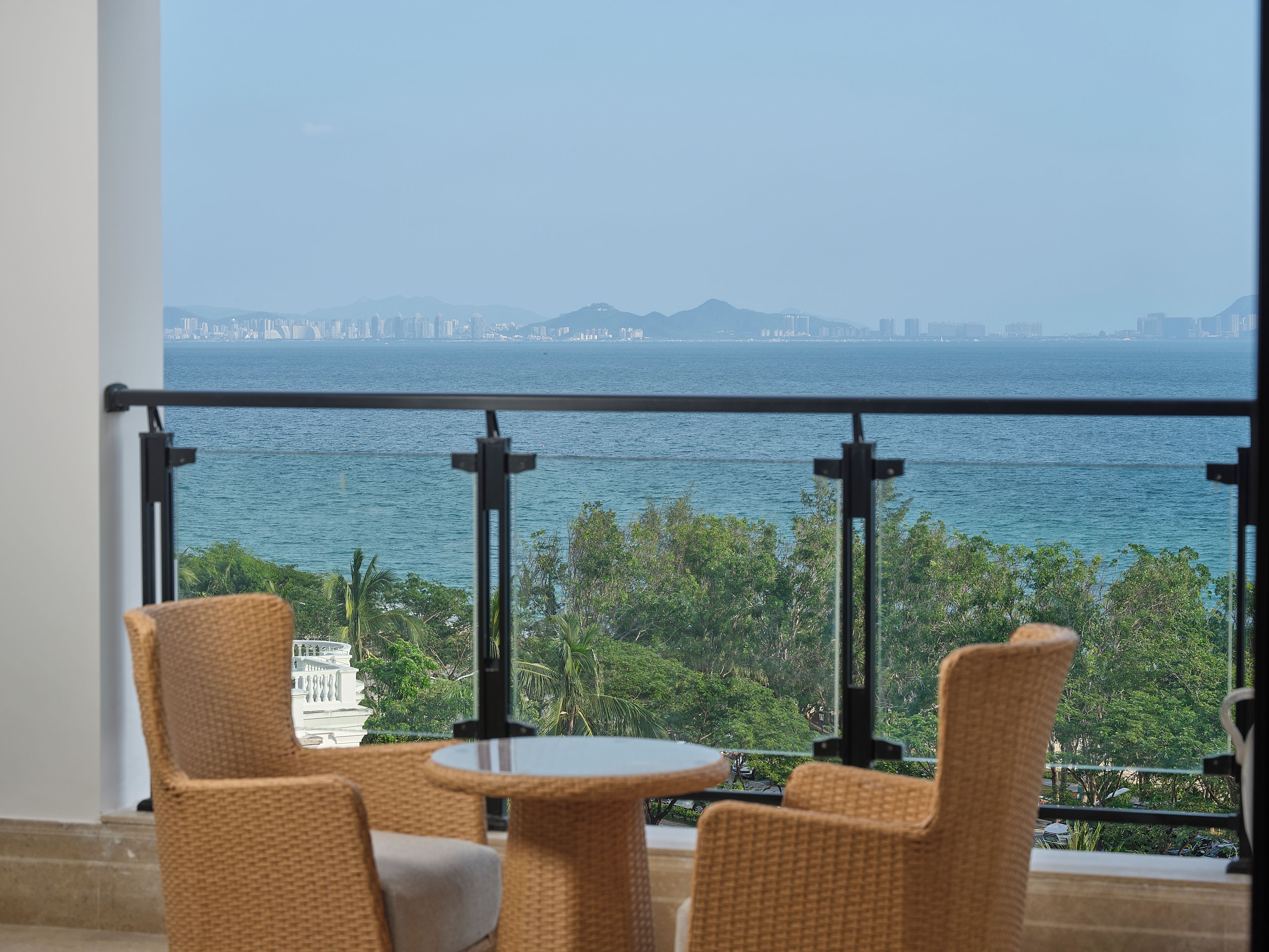 Ein Balkon mit Korbmöbeln und Blick auf das Meer sowie eine entfernte Stadtlandschaft.