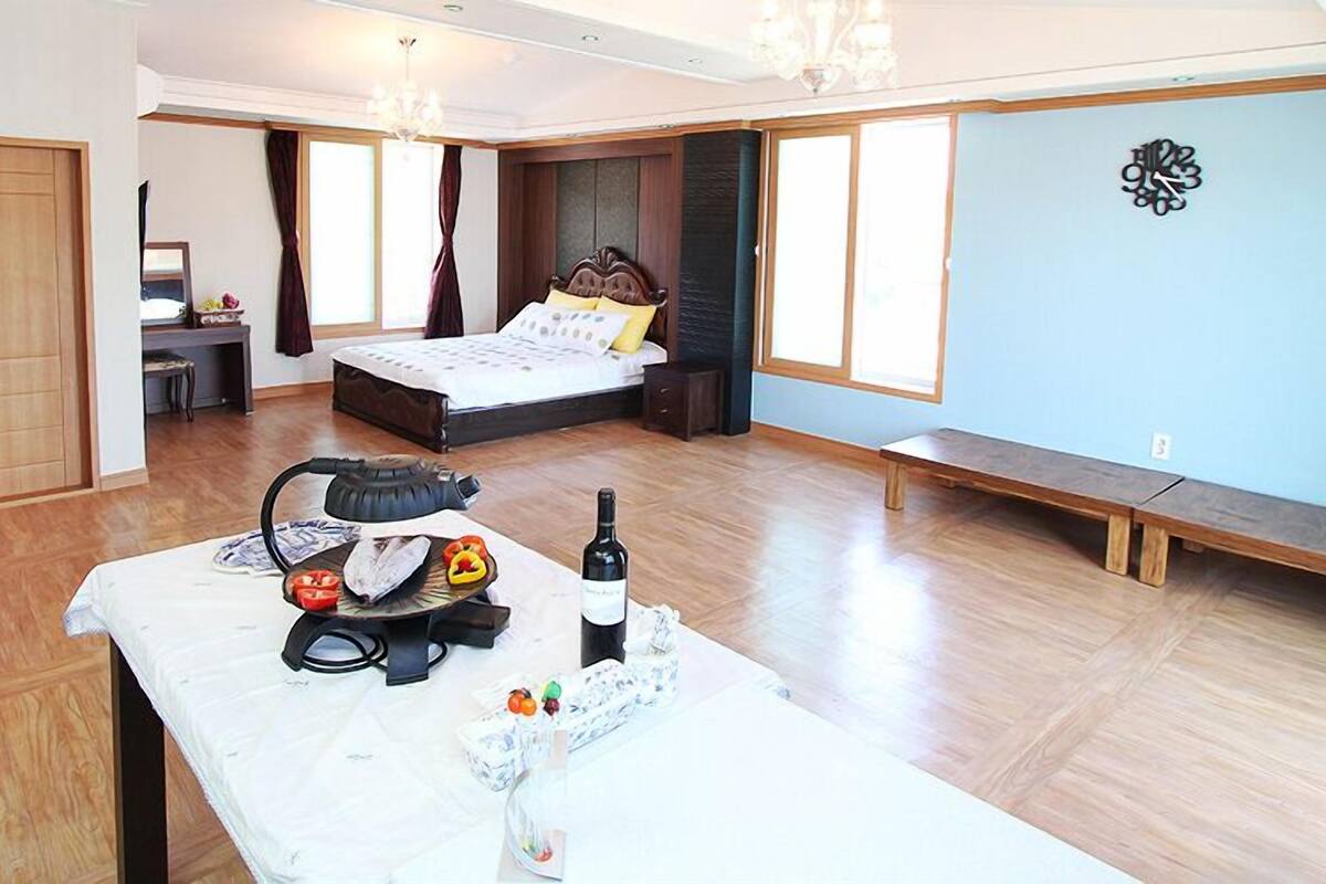 A spacious bedroom with a large bed, a wooden bench, a table with a kettle, and a bottle of wine.