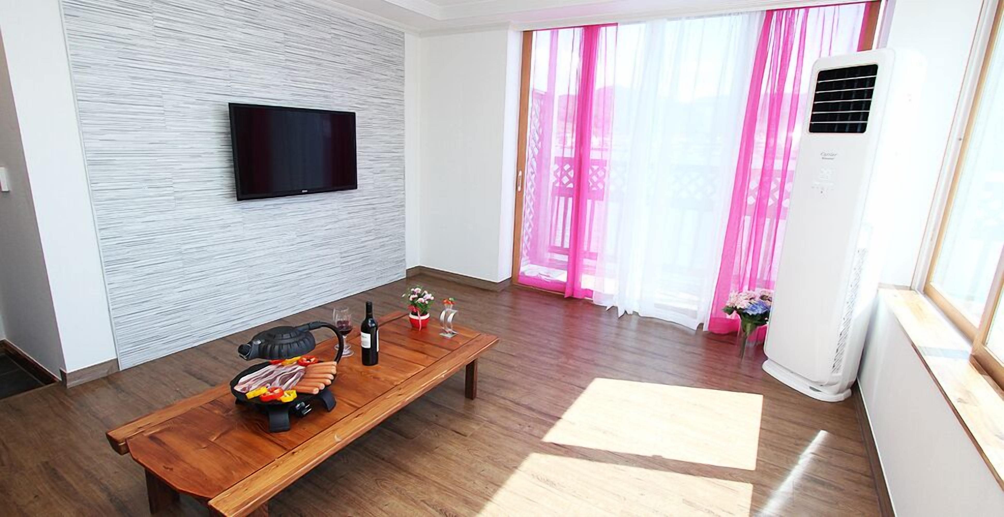 Una sala de estar moderna con un televisor de pantalla plana montado en una pared texturizada, una mesa de centro de madera con una botella de vino y flores, y un ventanal con cortinas rosadas.