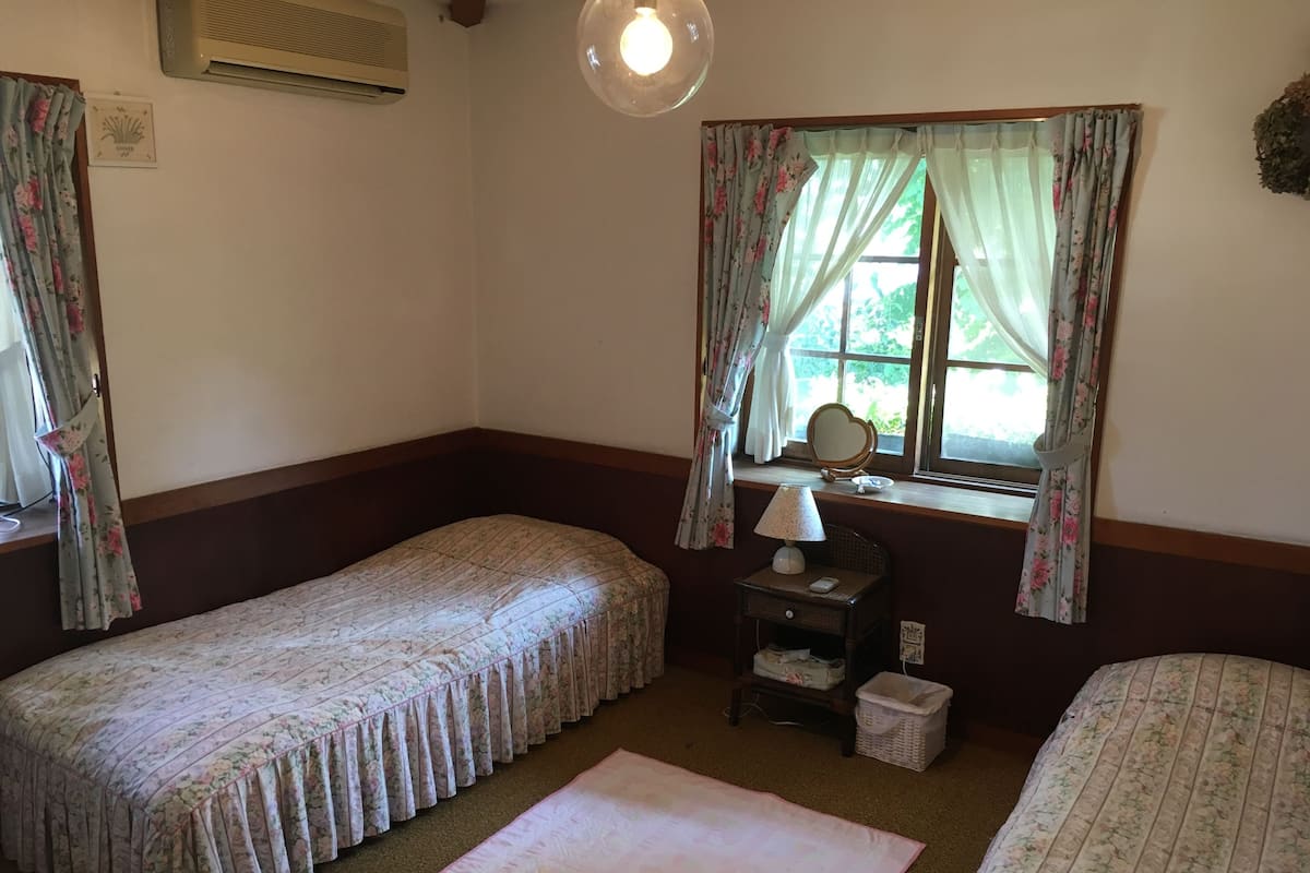 Un dormitorio con dos camas individuales, una mesita de noche con lámpara, una ventana con cortinas y un aire acondicionado de pared.