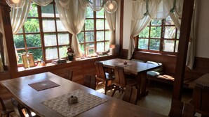 A dining area with wooden tables and chairs, large windows with curtains, and a view of greenery outside. - Pension Cocotte (Takayama)